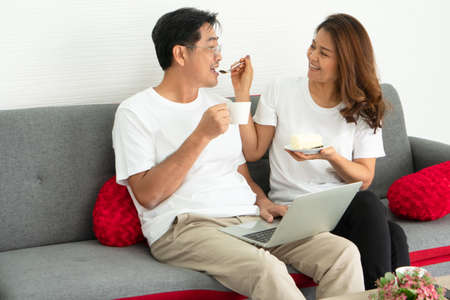 happy family Asian on vacation. Loving mature wife feeding middle-aged husband in the living room. senior couple retired stay home healthy talking smiling cheerful think positive optimism.の写真素材