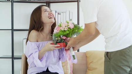 spouse in living room happy Asian couple with bunch flowers and gift at home. Young man is presenting flowers to his beloved Beautiful woman smiling while sitting on sofa. Surprise!  Valentine's Day.の写真素材