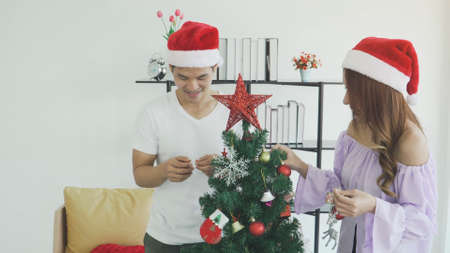happy smiling couple wear Santa hat decorating Christmas tree at home. Beautiful young woman and man...