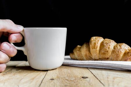 Man holding coffee cup with fresh croissant on white fabric. Wooden table and black background with copy spaceの写真素材