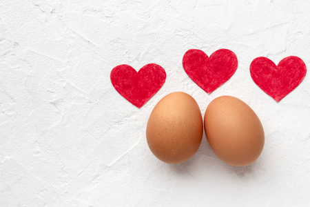 Two fresh eggs with three red paper hearts lie on white background with egg's shadowの写真素材