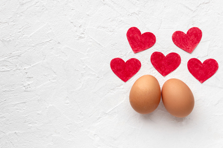Two fresh eggs with five red loving paper hearts lie on white textural background with egg's shadow Valentines day concept with copy spaceの写真素材