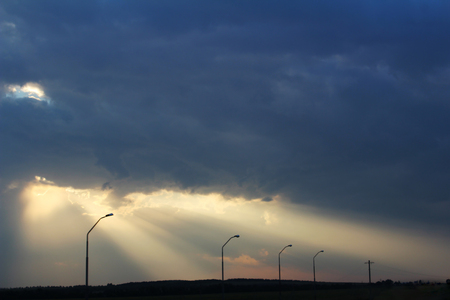 street lights in a row against the sky in front of a thunderstorm from which a ray of sunlight breaks throughの写真素材