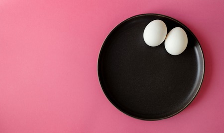 two white eggs lay on a dark plate, on a bright pink color background, top view with copy space