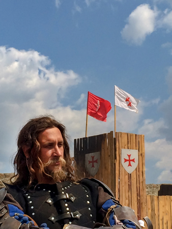 Sudak, Russia - August 16, 2015: brutal man with a beard, long hair and a stern look in the armor of a medieval knight against a wooden wall with flags and a coat of arms of the Crusadersのeditorial素材