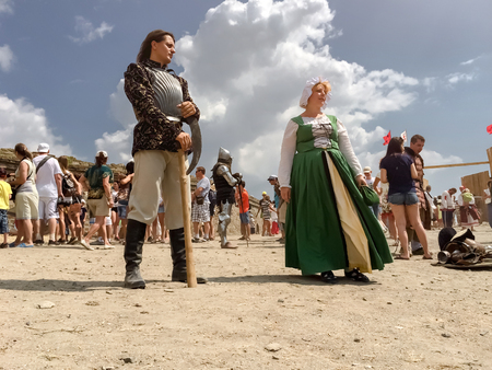 Sudak, Russia - August 16, 2015: rich medieval knight in steel armor holding an ax and a young lady maid in a green dress, corset and cap stand on the square, against the sky, the crowd and the castle, reconstruction of history, view from the ground levelのeditorial素材