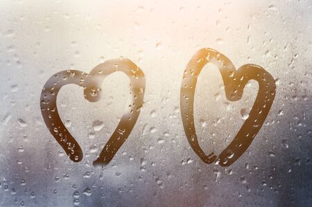 two hearts painted on glass in Rainy weather there are many drops on it and the sun shines outside the window at dusk between them, Valentine's Day conceptの写真素材