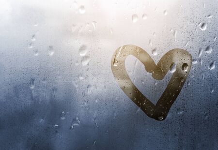 Heart painted on glass in Rainy weather, is fogged up and there are many drops on it, and the sun shines outside the window at dusk, toning in classic blueの写真素材