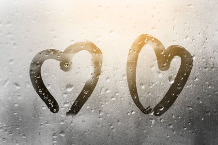 two hearts painted on glass in Rainy weather there are many drops on it and the sun shines outside the window at dusk between themの写真素材