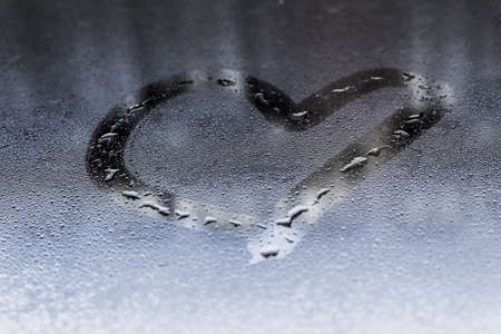 Heart painted on glass in Rainy weather, is fogged up and there are many drops on it, and the sun shines outside the window at dusk, toning in classic blueの写真素材