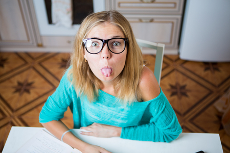Young woman sits at the table, looks at the canera and shows tongueの写真素材