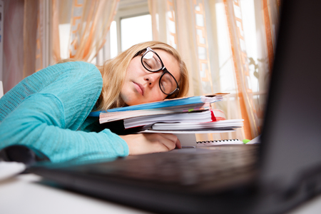 Young woman with glasses sleeping on pile of books next to laptopの写真素材