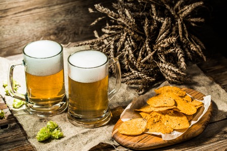 Two beer mugs with hops and chips and wheat on linen cloth on tableの写真素材