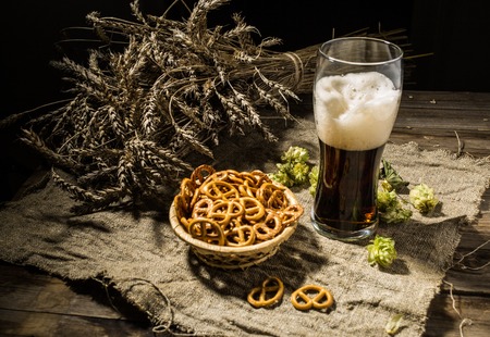 Glasse beer with wheat and hops, basket of pretzels on linen cloth on wooden tableの写真素材