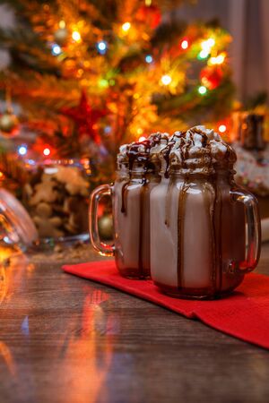 Two glasses of cocoa with whipped cream and chocolate syrup on wooden table and Gingerbread house, cookie jar and christmas tree with toys and garland on background. Christmas illustration.の写真素材