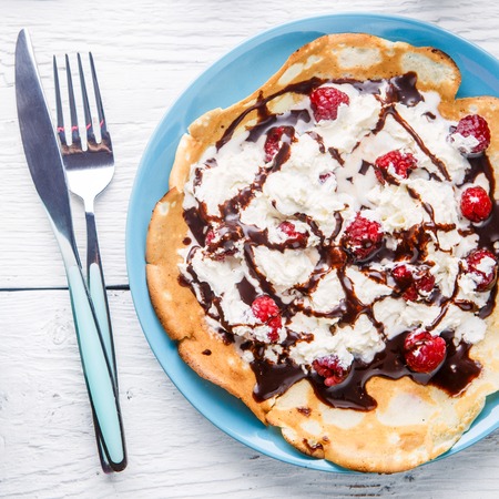 Homemade pancakes or Russian pancakes with chocolate sauce, whipped cream and raspberries on a plate on a white wooden background.の写真素材