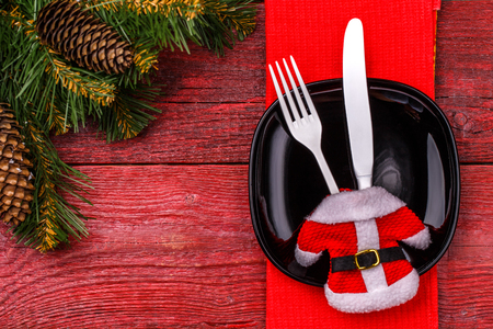 Christmas table place setting with red napkin, black plate, white fork and knife, decorated Santa jacket and christmas pine branches. Christmas holidays background.の写真素材