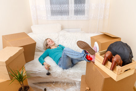 Photo of tired girl with glass of wine lying on sofa among cardboard boxesの写真素材