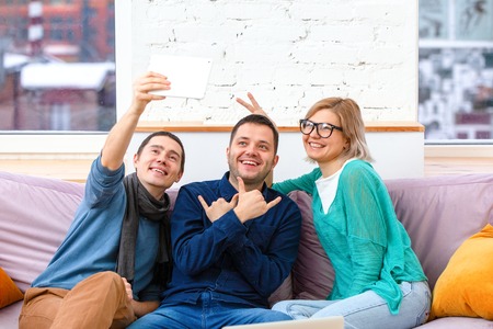 Friends sit on the couch, chatting and using a smartphone, desktop computer, laptop.の写真素材