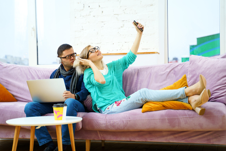 Man and woman sit on the couch, chatting and using smartphone, desktop computer.の写真素材