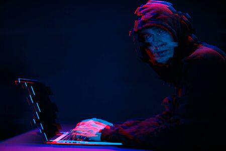 Hacker in the hood working with laptop typing text in dark room. Image with glitch effect