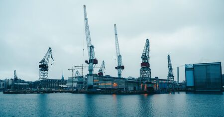 Photo of river, floating cranes, buildings on river bank in evening