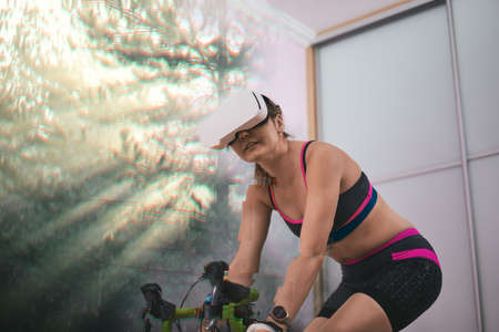 Woman trains at home on a stationary bike wearing VR-headset.の写真素材