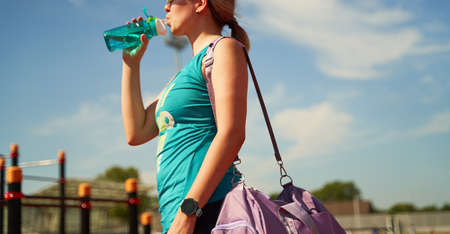 Female athlete with violet bag on her shoulder and water bottle stands near stadium waiting for...