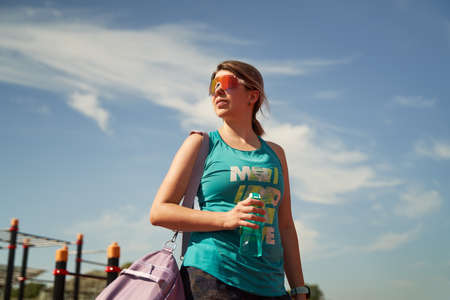 Female athlete with violet bag on her shoulder and water bottle stands near stadium waiting for training.の写真素材