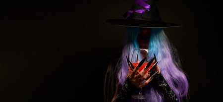 Witch with blue and violet hair, black hat with violet ribbon and long black nails is holding wine glass in her hands.の写真素材