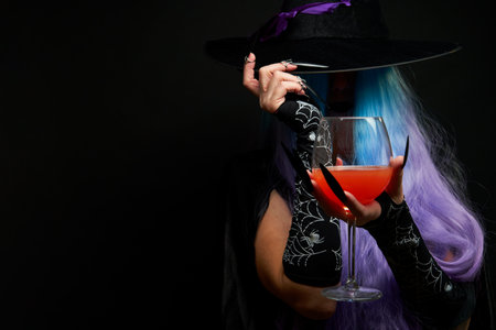 Witch with blue and violet hair, black hat with violet ribbon and long black nails is holding wine glass in her hands.の写真素材