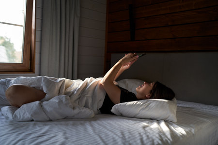 Woman in bed checking social media with smartphoneの写真素材