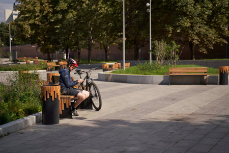 Man in a bicycle helmet sits on a bench and looks at his phone, resting while cycling in the cityの写真素材