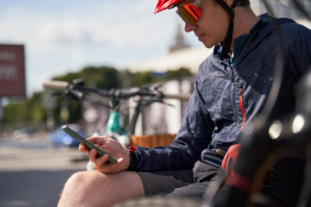 Man in a bicycle helmet sits on a bench and looks at his phone, resting while cycling in the cityの写真素材