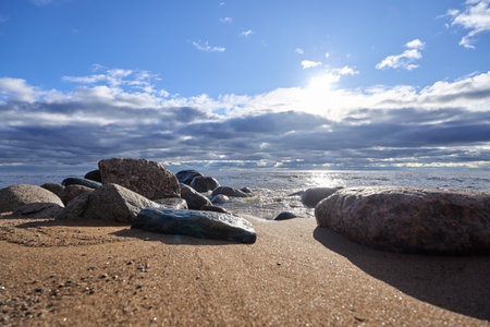Rocks on sandy shore with sunlight reflection and cloudy sky above sea.の写真素材