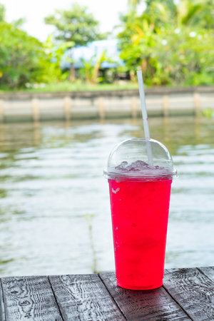 Red juice with ice in a plastic glass on table and river.の写真素材