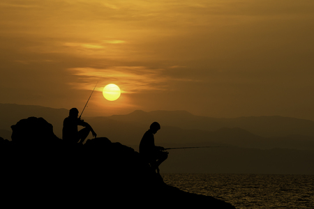Silhouette of tourists Fishing on the rocks in sea and Golden light of sunrise behind the mountains.の写真素材