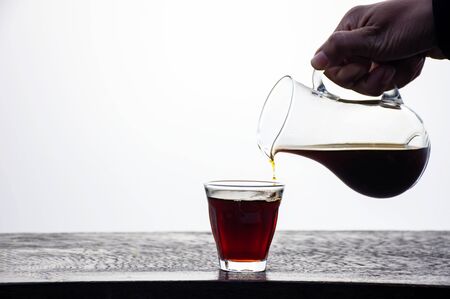 Hand holding a jug of black coffee poured into a glass on wooden table.の写真素材