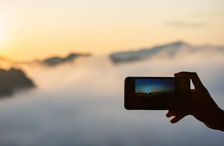 Hand holding the phone shooting the morning sun and the fog.の写真素材