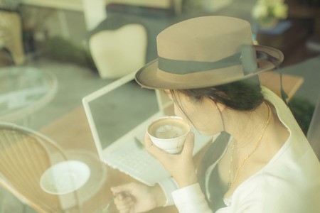 Asian woman drinking coffee and using computer in cafe. Photo filter effect.の写真素材