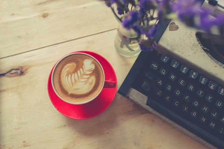 Red latte coffee cup with purple flower and typewriter on wooden background.