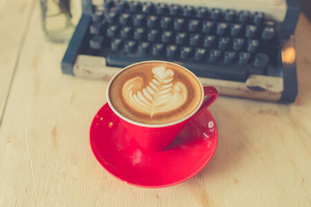 Red latte coffee cup with purple flower and typewriter on wooden background.