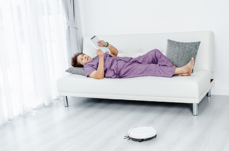 Senior woman unsing tablet while robot vacuum cleaning floor at home. Modern lifestyle concept.の写真素材