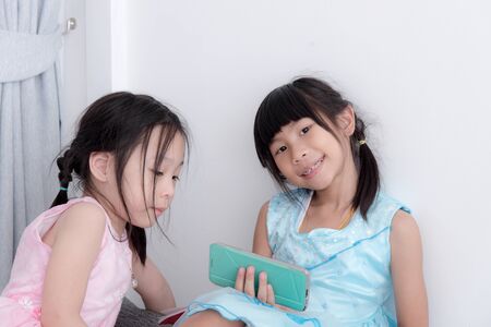 Two Asian girls playing with smart phone and sitting near white wall.の写真素材