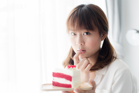 Cute Asian teenager holding strawberry cake and licking her finger at home.の写真素材