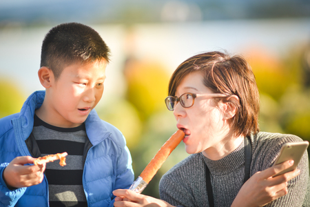 Asian boy feeding sausage to his Aunt while she is using smart phone outdoor.の写真素材