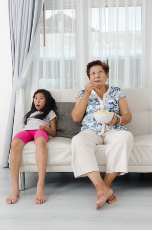 Asian senior woman eating popcorn with her grandchild while watching TV at home, selective focusの写真素材