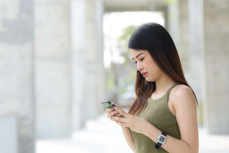 Young Asian woman with smartphone standing against street blurred building background.