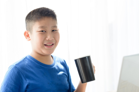 Happy Asian boy holding black cup while using tablet at home.の写真素材