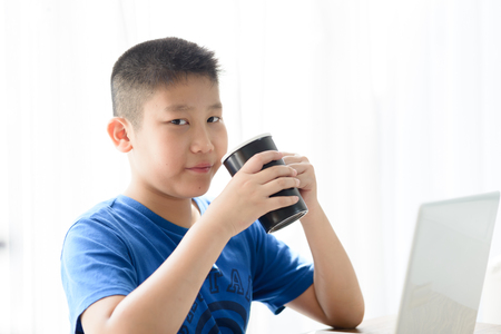 Happy Asian boy holding black cup while using tablet at home.の写真素材
